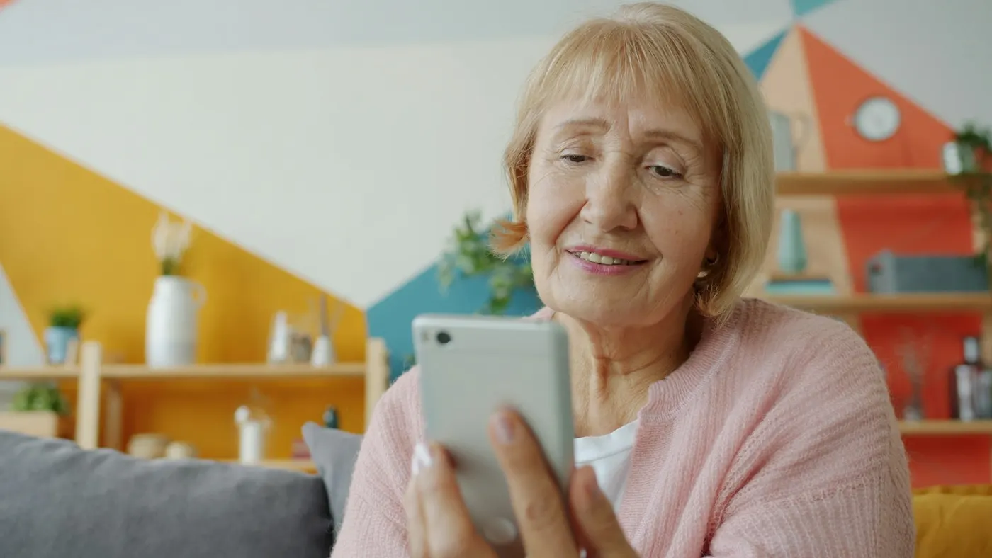 Une femme d’une soixantaine d’années sourit en lisant un message sur son téléphone, dans son salon